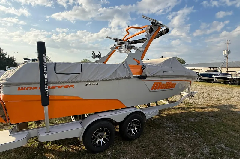 Slide: The Image of 2023 Malibu Wakesetter 23 LSV boat on trailer, covered, with orange accents, under a partly cloudy sky. - 12