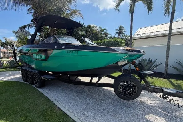 Slide: The Image of 2021 Malibu M240 boat on trailer, parked in tropical setting with palm trees. - 2