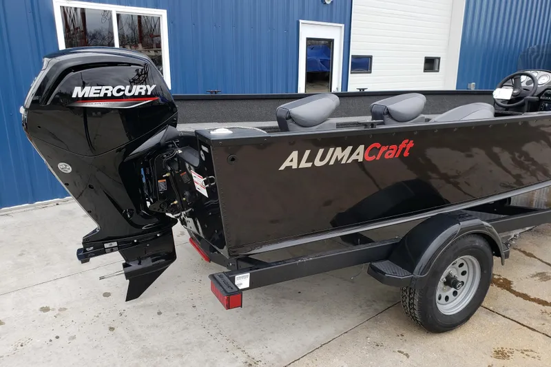 Slide: The Image of 2026 Alumacraft Voyageur 175 SC boat with Mercury outboard motor, parked near blue building. - 5
