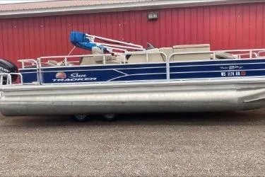 The Image of 2015 Sun Tracker Fishin' Barge 22 DLX pontoon boat, blue and silver, parked by a red building. - 1