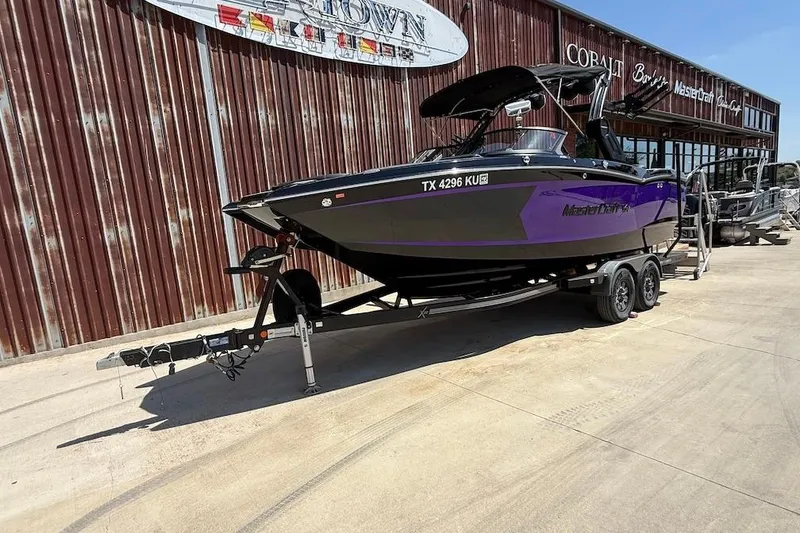Slide: The Image of 2023 MasterCraft XStar S Piranha boat on trailer, parked outside a rustic building. - 1