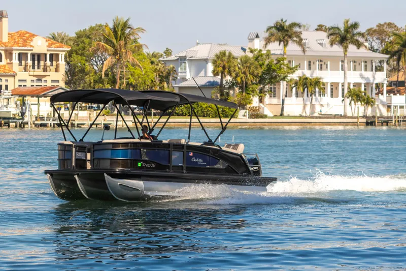 Slide: The Image of 2024 Barletta L23U pontoon boat cruising on a scenic waterfront with luxury homes. - 8