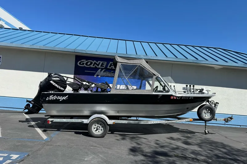 The Image of Jetcraft 1925 Adventurer boat on trailer, parked outside a building, under clear blue sky. - 1