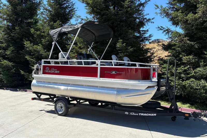 The Image of 2026 Sun Tracker Bass Buggy 16 XL Select pontoon boat on trailer, parked outdoors. - 0