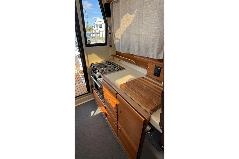 Slide: The Image of 2024 Ranger Tugs R-27 interior kitchen with stove, wooden cabinetry, and window view. - 23