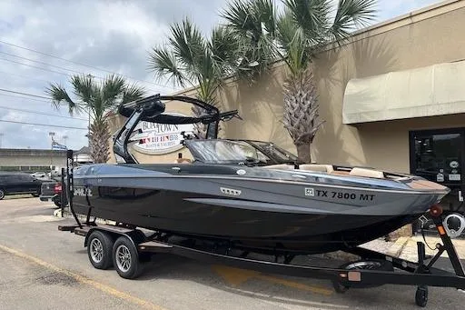 Slide: The Image of 2024 Supreme S220 boat on trailer, parked near palm trees and a building. - 2