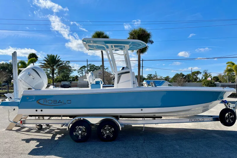 The Image of 2026 Robalo 246 Cayman boat on trailer, blue hull, sunny day, palm trees in background. - 1