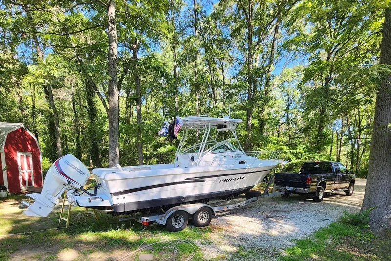 The Image of 1999 Pro-Line 241 Walkaround boat on trailer, parked in wooded area with truck nearby. - 0