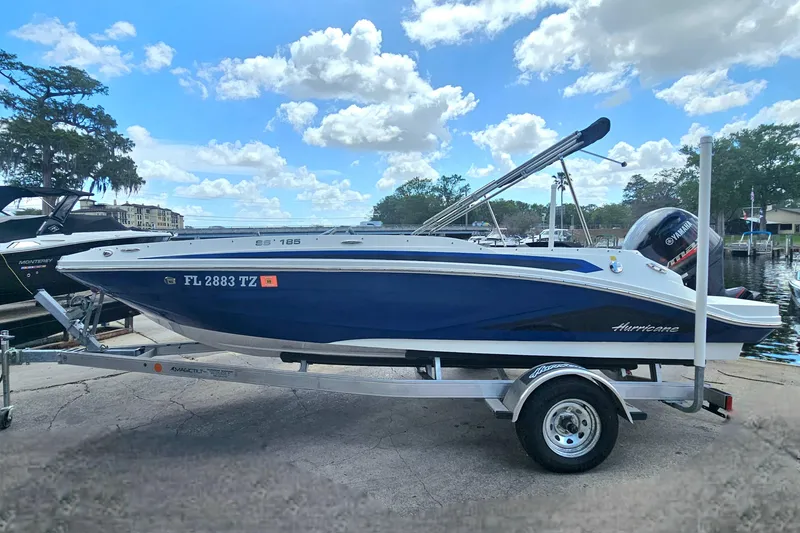 Slide: The Image of 2023 Hurricane SunDeck Sport 188 OB boat on trailer, parked near a marina under a blue sky. - 5