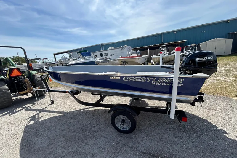 Slide: The Image of 2026 Crestliner 1469 Outreach boat on trailer, parked outdoors near a blue building. - 2