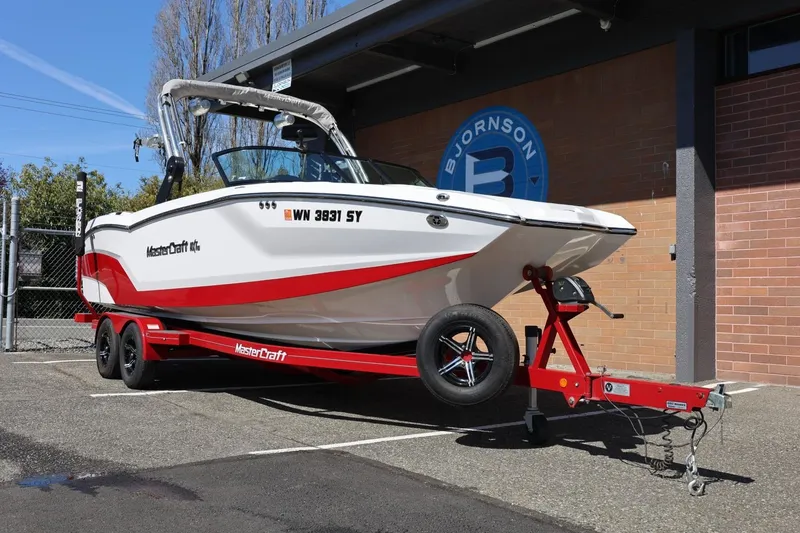 Slide: The Image of 2021 MasterCraft NXT24 boat on red trailer, parked outdoors near brick building. - 4