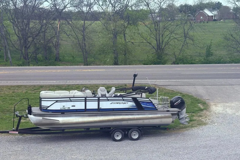 The Image of 2023 Starcraft SLS 3 pontoon boat on trailer, parked near a road with trees in the background. - 0