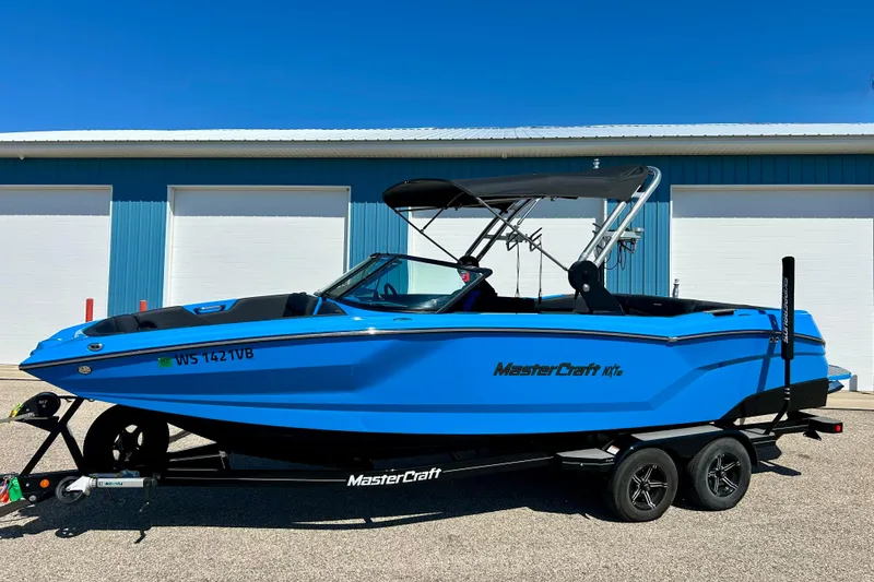 Slide: The Image of 2021 MasterCraft NXT24 boat in vibrant blue, parked on a trailer, against a blue building. - 3