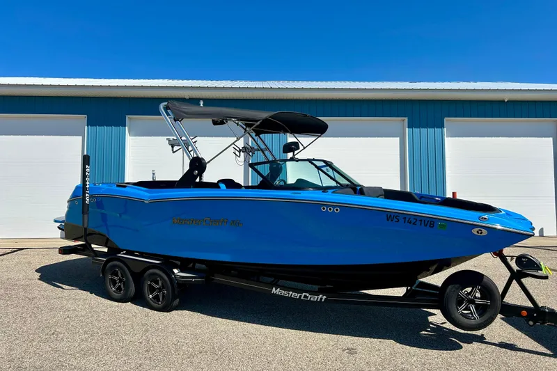 Slide: The Image of 2021 MasterCraft NXT24 boat in vibrant blue, parked on a trailer, against a blue building. - 6