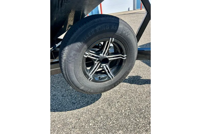 Slide: The Image of 2021 MasterCraft NXT24 trailer tire on asphalt, showcasing black alloy wheel design. - 67
