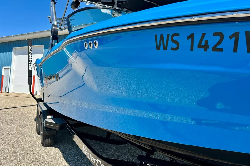 Slide: The Image of 2021 MasterCraft NXT24 boat in vibrant blue, parked on a trailer near a blue building. - 9