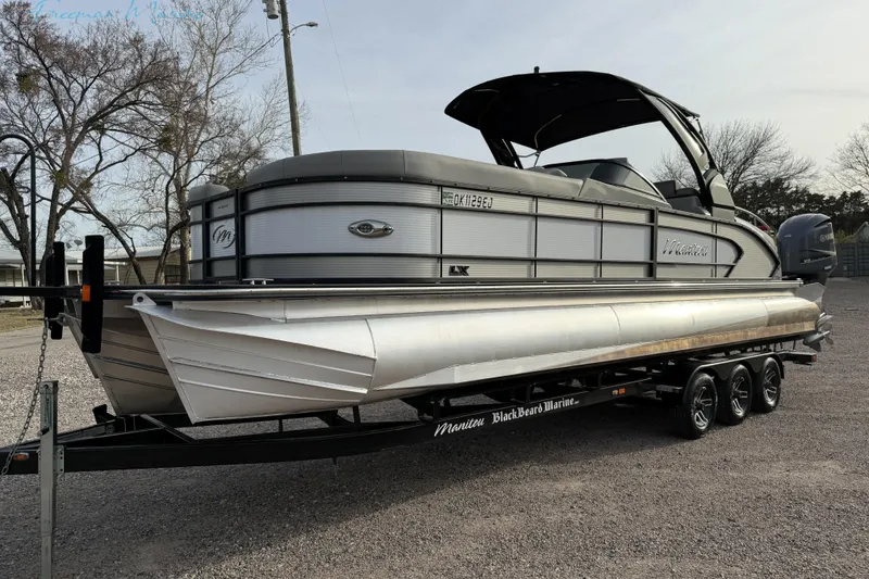 Slide: The Image of 2022 Manitou 27 LX SRW pontoon boat on a trailer, parked outdoors. - 1