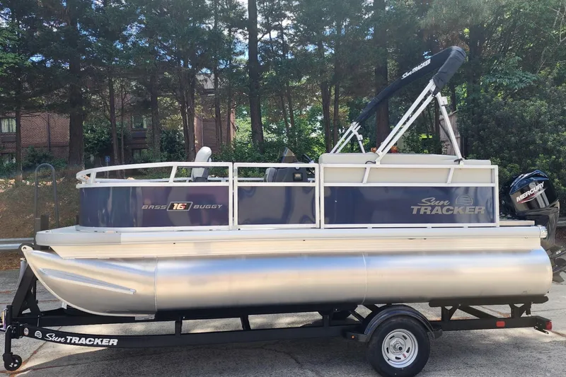 The Image of 2026 Sun Tracker Bass Buggy 16 XL Select pontoon boat on trailer, parked outdoors. - 0