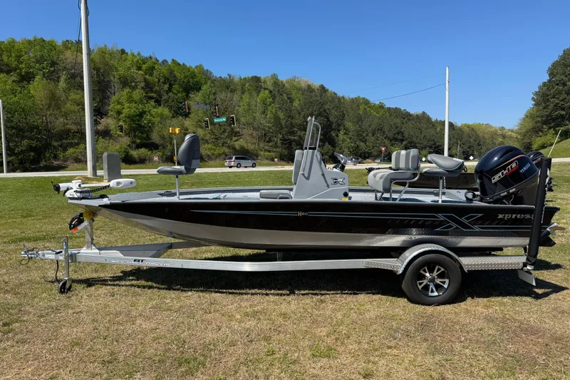 The Image of 2025 Xpress H20B Bay boat on trailer, parked on grass, with trees and road in background. - 0