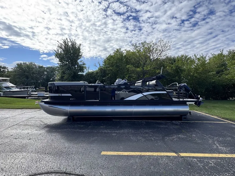 The Image of A 2026 Sylvan L3 DLZ pontoon boat is parked on a lot, surrounded by trees and a cloudy sky. - 0