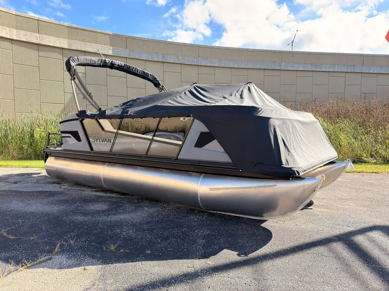 Slide: The Image of A 2027 Sylvan R-20 Q DH pontoon boat with a black cover, parked on a paved area. - 1