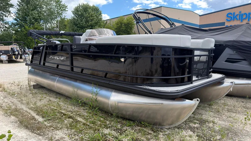 Slide: The Image of A modern pontoon boat with black railing and silver pontoons, parked on a gravel lot. - 1