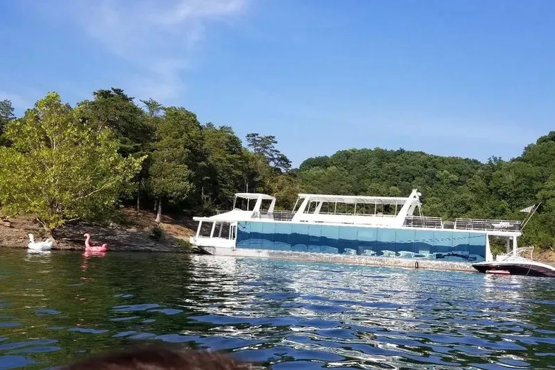 The Image of 2012 Thoroughbred Houseboat 21x103 on a scenic lake with lush greenery. - 0