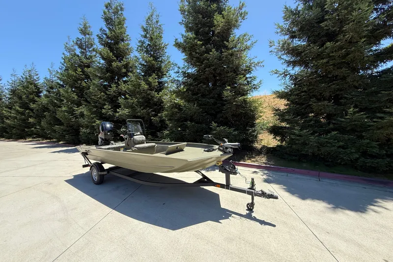 Slide: The Image of 2026 Tracker Grizzly 1860 CC boat on trailer, parked near tall trees under clear blue sky. - 2
