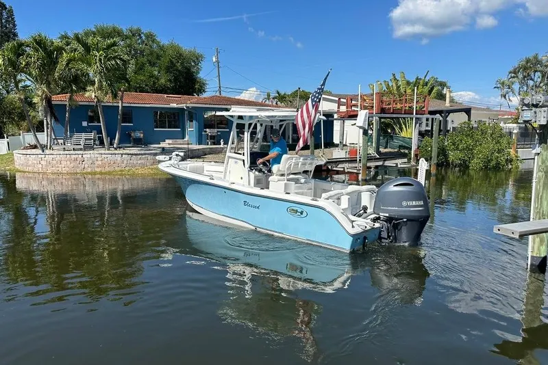 Slide: The Image of 2018 Sea Hunt Ultra 225 boat on calm water near a dock, with a house and palm trees. - 18