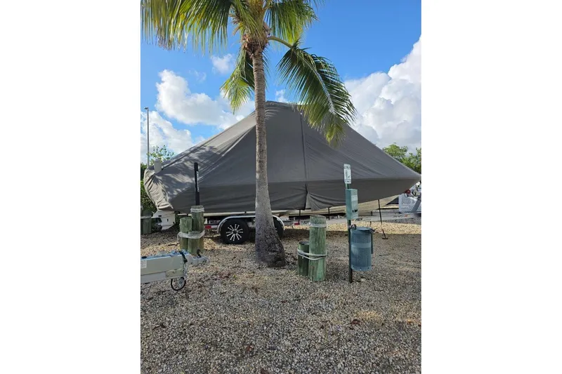 Slide: The Image of 2024 Pathfinder 2400 Open boat covered, parked under a palm tree on gravel. - 2