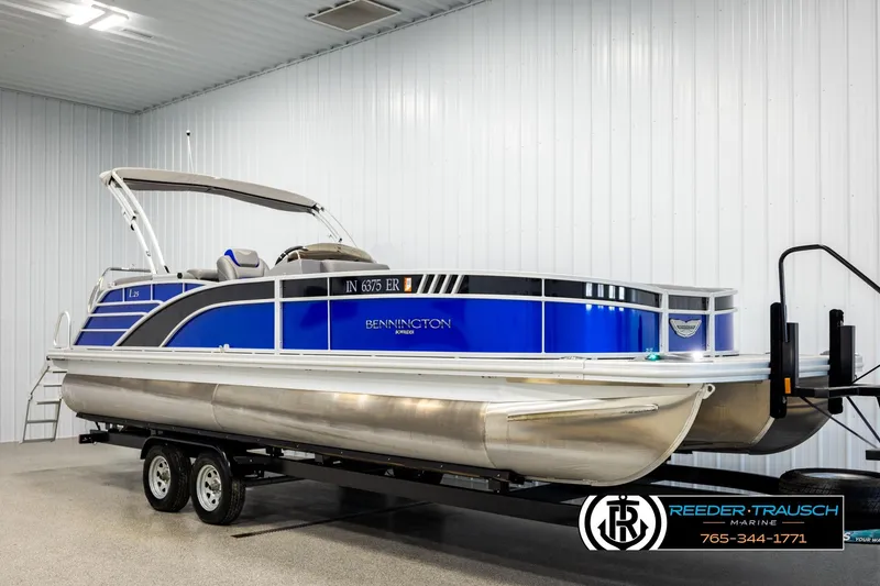 Slide: The Image of 2025 Bennington LTFBSE pontoon boat in showroom, featuring sleek blue design on trailer. - 2