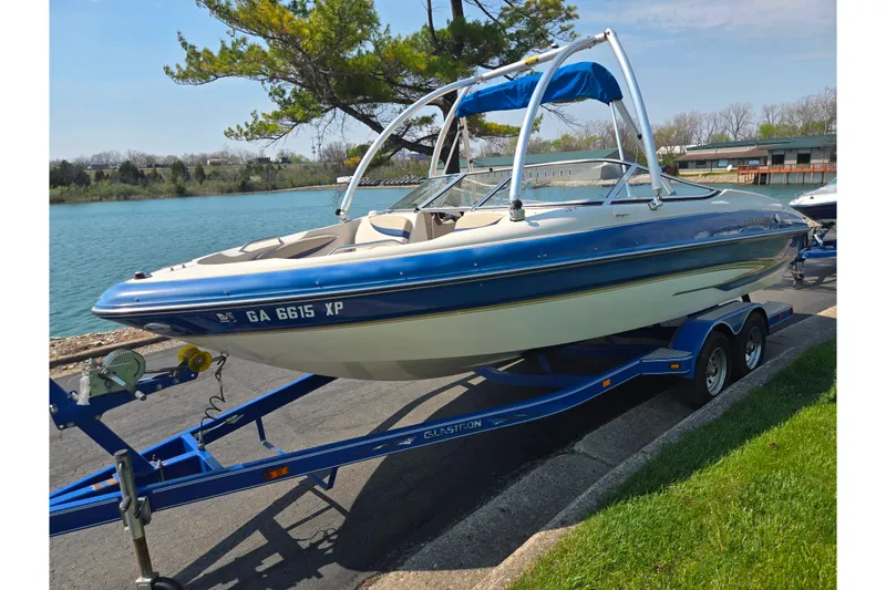 Slide: The Image of 2005 Glastron GX 235 boat on trailer by a lake, featuring blue and white design. - 9