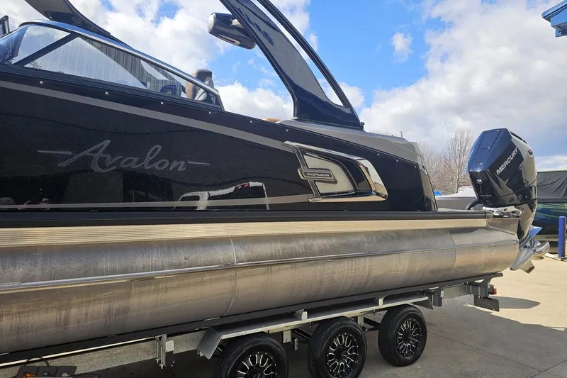 Slide: The Image of 2024 Avalon Excalibur Quad Lounge Windshield boat on trailer, featuring sleek design and Mercury engine. - 27
