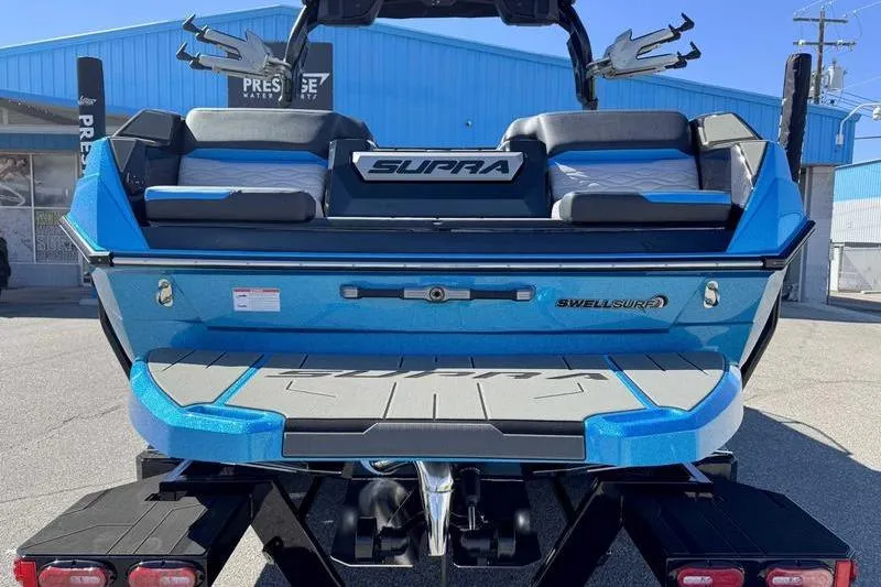 Slide: The Image of 2026 Supra SL boat with blue exterior, featuring Swell Surf system, parked outdoors. - 2