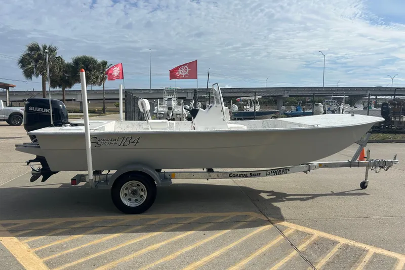 Slide: The Image of 2026 Coastal Skiff 184 boat on trailer, parked outdoors with flags and palm trees. - 4