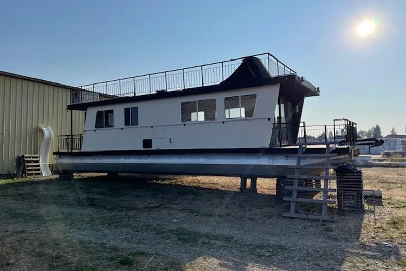 The Image of 1985 Three Buoys houseboat on land, with upper deck and slide, under sunny sky. - 0