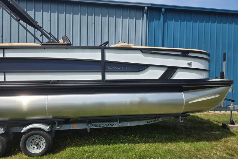 Slide: The Image of 2026 Evotti 523QL pontoon boat on trailer, parked beside blue building. - 2