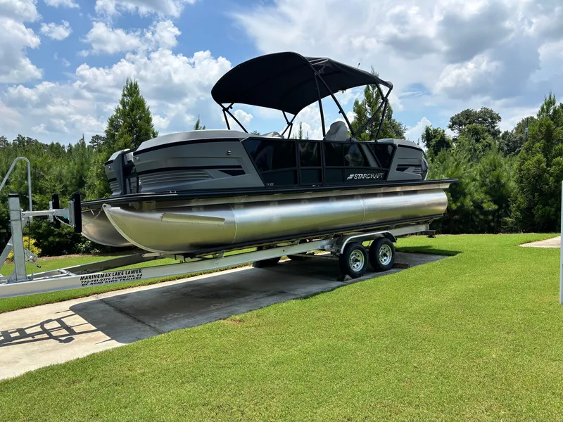 Slide: The Image of A 2026 Starcraft VX 22 R DH pontoon boat on a trailer, set on green grass. - 0