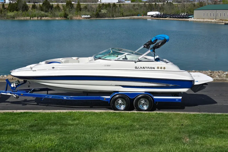 The Image of 2006 Glastron DX 235 boat on trailer by a lake, showcasing sleek design and blue accents. - 0