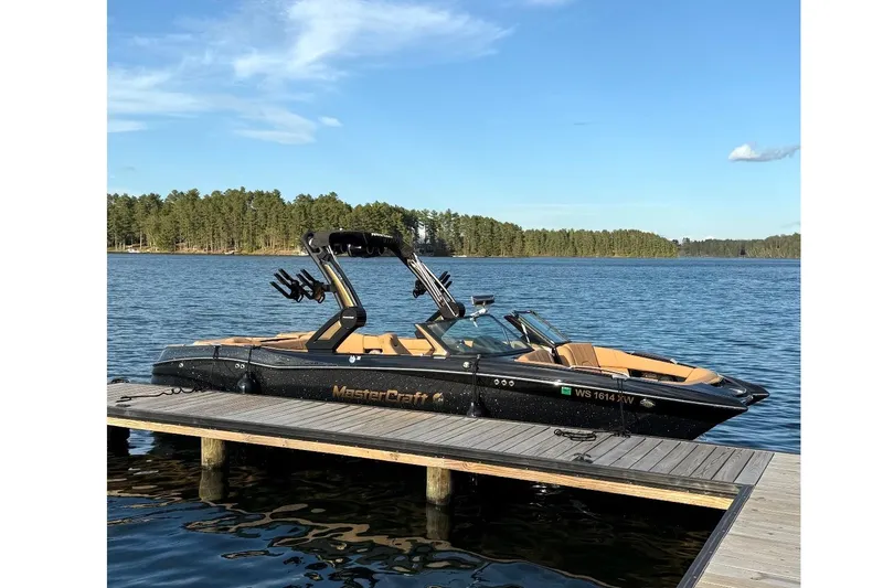 The Image of 2024 MasterCraft X24 boat docked on a serene lake with forested background. - 1