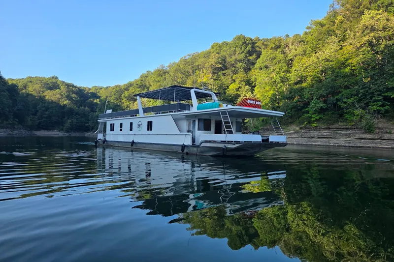 The Image of 2003 Fantasy 18x80 houseboat on a serene lake surrounded by lush green trees. - 0