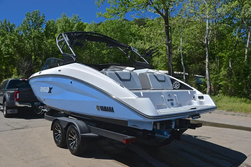 Slide: The Image of 2024 Yamaha Boats 252XE on trailer, parked near lush green trees. - 12