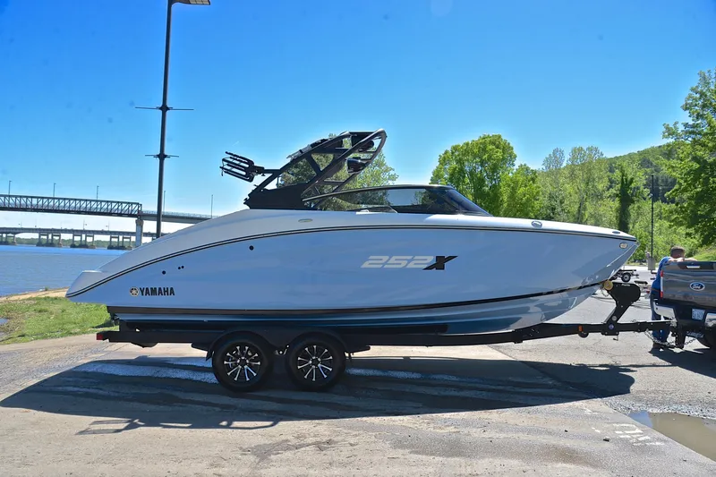Slide: The Image of 2024 Yamaha Boats 252XE on trailer by riverside, sunny day, bridge in background. - 15