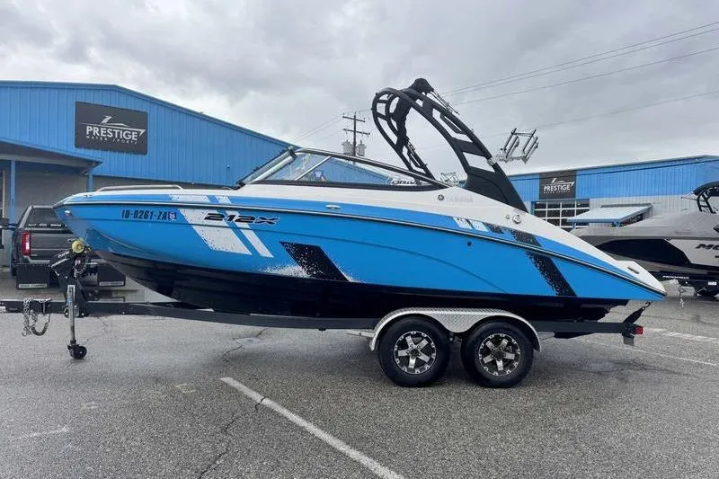 The Image of 2021 Yamaha Boats 212X in blue, parked on a trailer outside a dealership. - 1