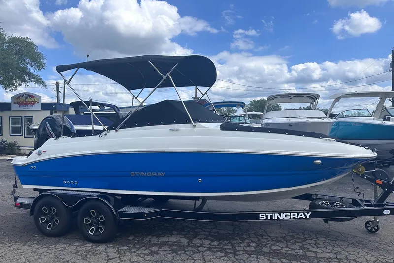 The Image of 2022 Stingray 201 DC boat with blue hull on trailer, parked outdoors under cloudy sky. - 0