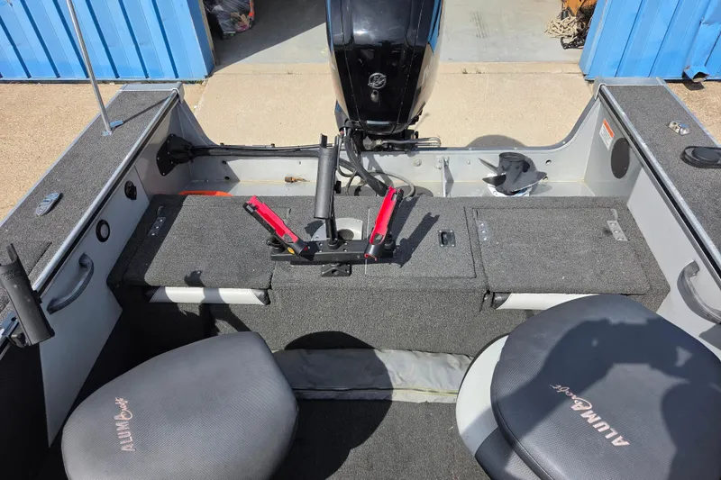 Slide: The Image of 2016 Alumacraft Trophy 175 boat interior with seating and motor, docked near blue building. - 9