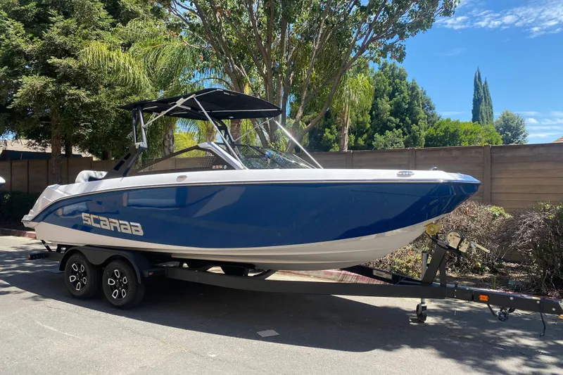 The Image of 2025 Scarab 235 SE boat on trailer, parked outdoors, surrounded by trees and a wooden fence. - 0