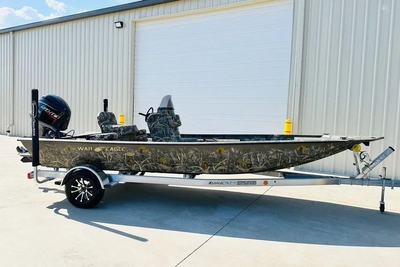 The Image of 2026 War Eagle 961 Blackhawk boat on trailer, camo design, parked outside a warehouse. - 1