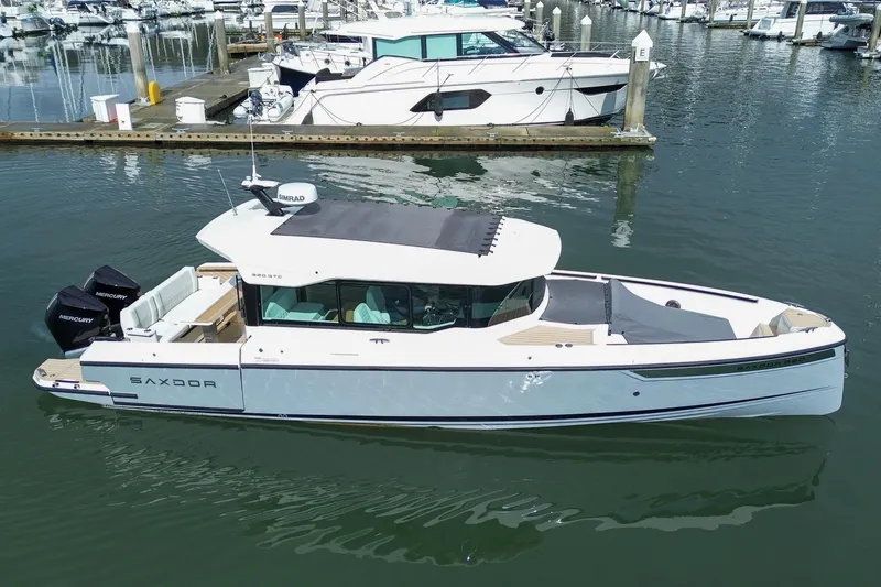 The Image of 2026 Saxdor 320 GTC boat docked in marina, featuring sleek design and twin Mercury engines. - 0