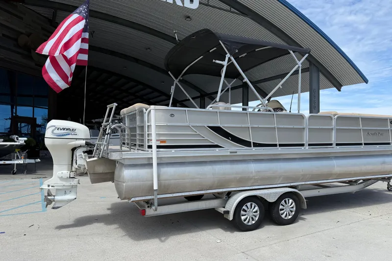 Slide: The Image of 2012 South Bay 522 CR pontoon boat on trailer, parked near a building with an American flag. - 3